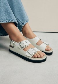 Sandali bianchi con due grandi fibbie argentate, tallone aperto e suola in gomma nera. Indossati con jeans a gamba larga azzurri.