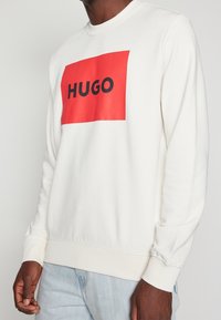 Sweatshirt bianco con polsini a costine, caratterizzato da un grande logo rettangolare rosso con la scritta "HUGO" in lettere nere sul petto.