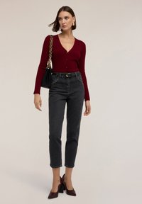 Cárdigan rojo rubí con textura de canalé, jeans negros de tiro alto, zapatos de tacón burdeos y un bolso negro de hombro con detalle de cadena dorada.