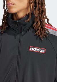 Chaqueta negra con cremallera y cuello alto, con rayas rojas y grises, parche del logo de Adidas y una tela suave y texturizada.