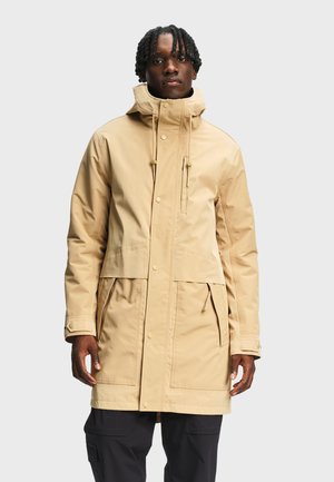 Homme portant un long parka beige à capuche avec plusieurs poches et un pantalon noir, debout devant un fond clair uni.