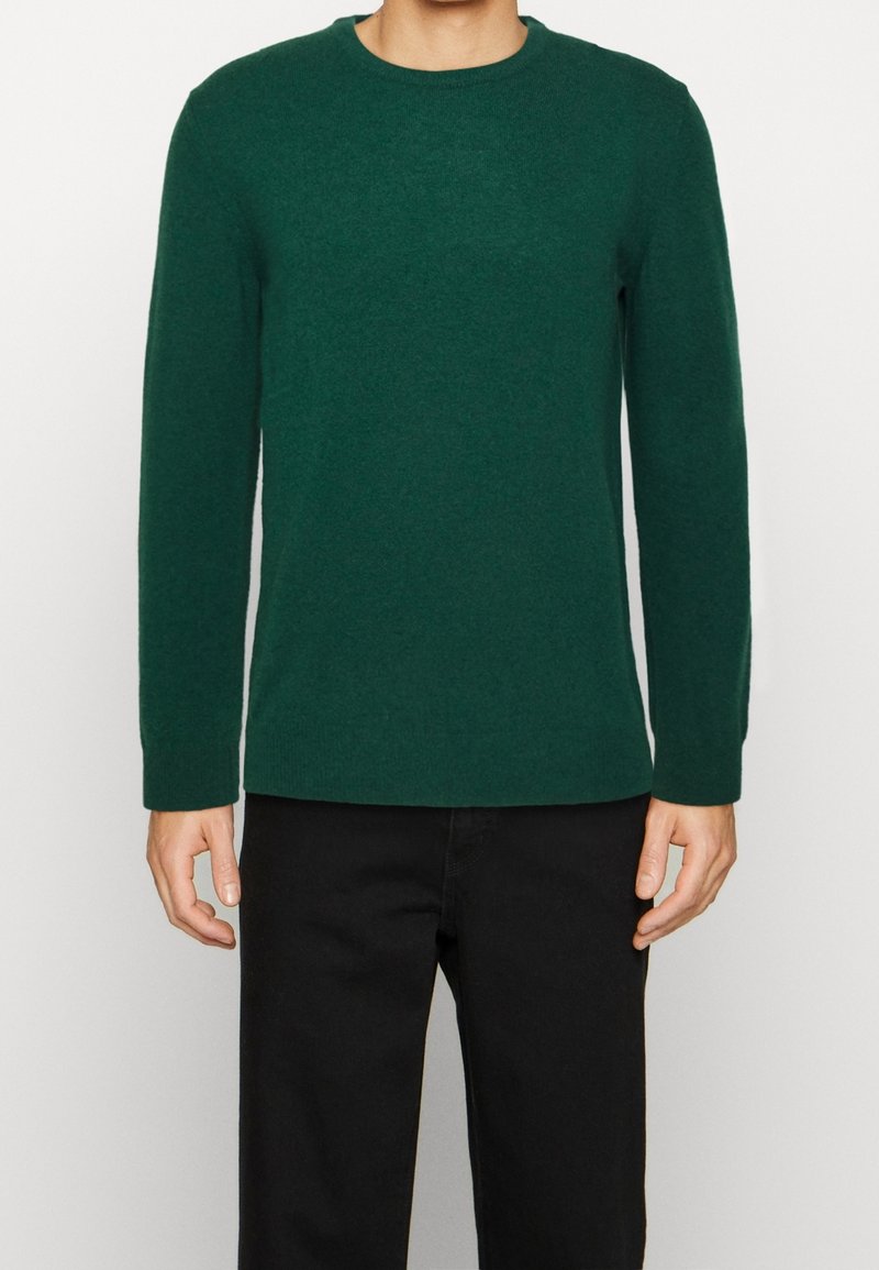 Homme portant un pull uni vert foncé à manches longues et un pantalon noir, debout avec les bras détendus sur un fond clair uni.