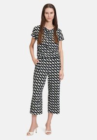 Granatowy kombinezon w falisty wzór w kolorze kremowym, z krótkimi rękawami, szerokimi, skróconymi nogawkami i bocznymi kieszeniami. Modelka nosi białe mule na obcasie.