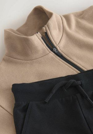 Kreminės spalvos zip-up tipo megztinis, pagamintas iš minkštos medžiagos, su aukštu apykakle ir juodomis detalėmis. Derinamas su juodomis sportinėmis kelnėmis, turinčiomis ripsinę juosmenį ir raištelį.