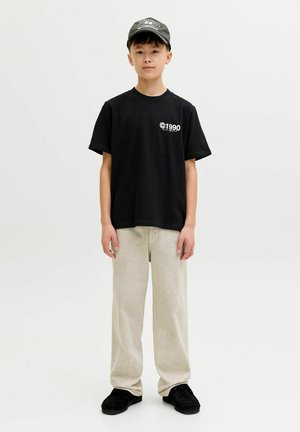 Czarny bawełniany t-shirt z białym nadrukiem "©1990", w połączeniu z jasnobeżowymi sztruksowymi spodniami i czarnymi zamszowymi butami. Na głowie ciemna czapka.