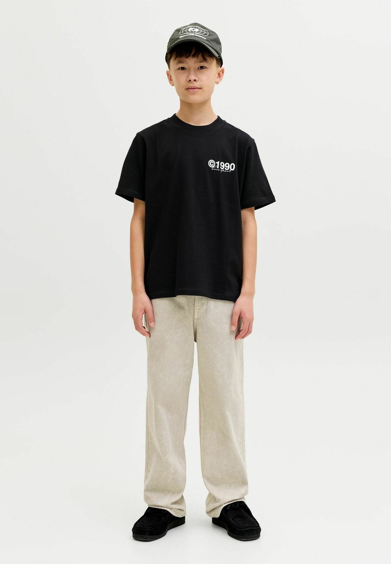 Czarny bawełniany t-shirt z białym nadrukiem "©1990", w połączeniu z jasnobeżowymi sztruksowymi spodniami i czarnymi zamszowymi butami. Na głowie ciemna czapka.