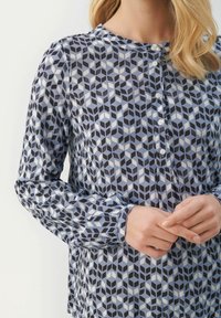 Camicia a maniche lunghe con motivo geometrico blu e nero, caratterizzata da colletto tondo e pattina dei bottoni. Tessuto liscio con un leggero lucido.