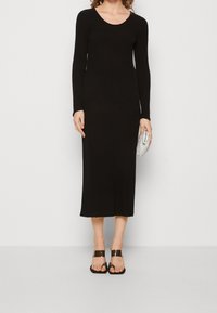 Robe maxi noire côtelée avec manches longues, encolure ronde et ourlet festonné. Portée avec des sandales noires et une pochette argentée.