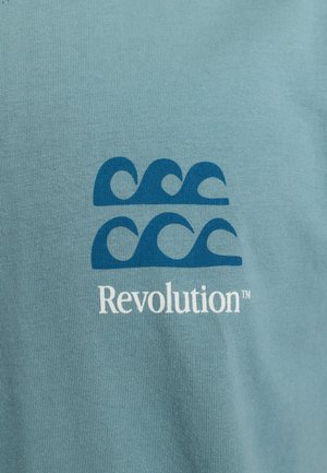 Lichtblauwe katoenen t-shirt met een grafische weergave van blauwe golfpatronen boven de tekst "Revolution™" in het wit. Zachte, gladde textuur.