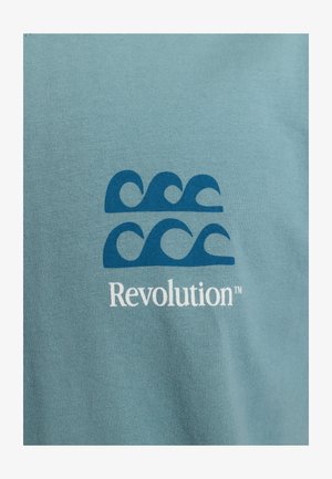 T-shirt en coton bleu clair featuring des motifs de vagues bleues au-dessus du texte "Revolution™" en blanc. Texture douce et lisse.