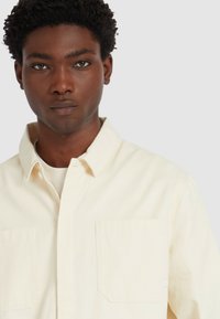 Młody mężczyzna z ciemnymi kręconymi włosami, ubrany w kremową koszulę zapinaną na guziki na białą koszulkę, patrzący na wprost z neutralnym wyrazem twarzy.