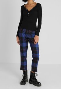 Haut à manches longues noir avec détail de boutons, associé à un pantalon à carreaux bleu et noir. Le mannequin porte des bottines à lacets noires.