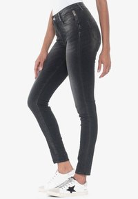 Jeans negros ajustados y skinny, confeccionados en una tela de denim suave con un acabado desgastado, que cuentan con cinco bolsillos y cierre de cremallera. Zapatillas blancas con un acento de estrella negra.