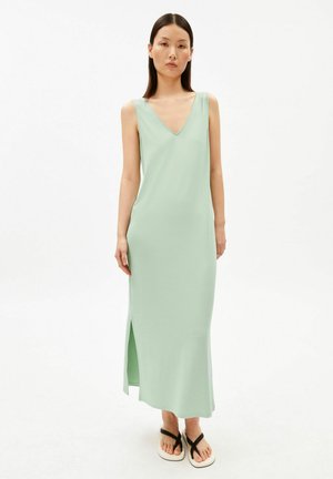 Femme portant une robe sans manches vert clair à encolure en V, longueur chevilles avec une fente sur le côté, et des sandales noires à lanières, debout devant un fond blanc.