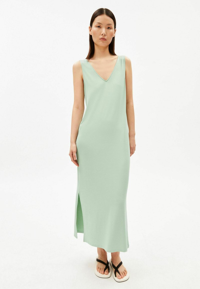 Frau trägt ein ärmelloses, hellgrünes, knöchellanges Kleid mit V-Ausschnitt, Seitenschlitz und schwarzen Riemchensandalen, steht vor weißem Hintergrund.