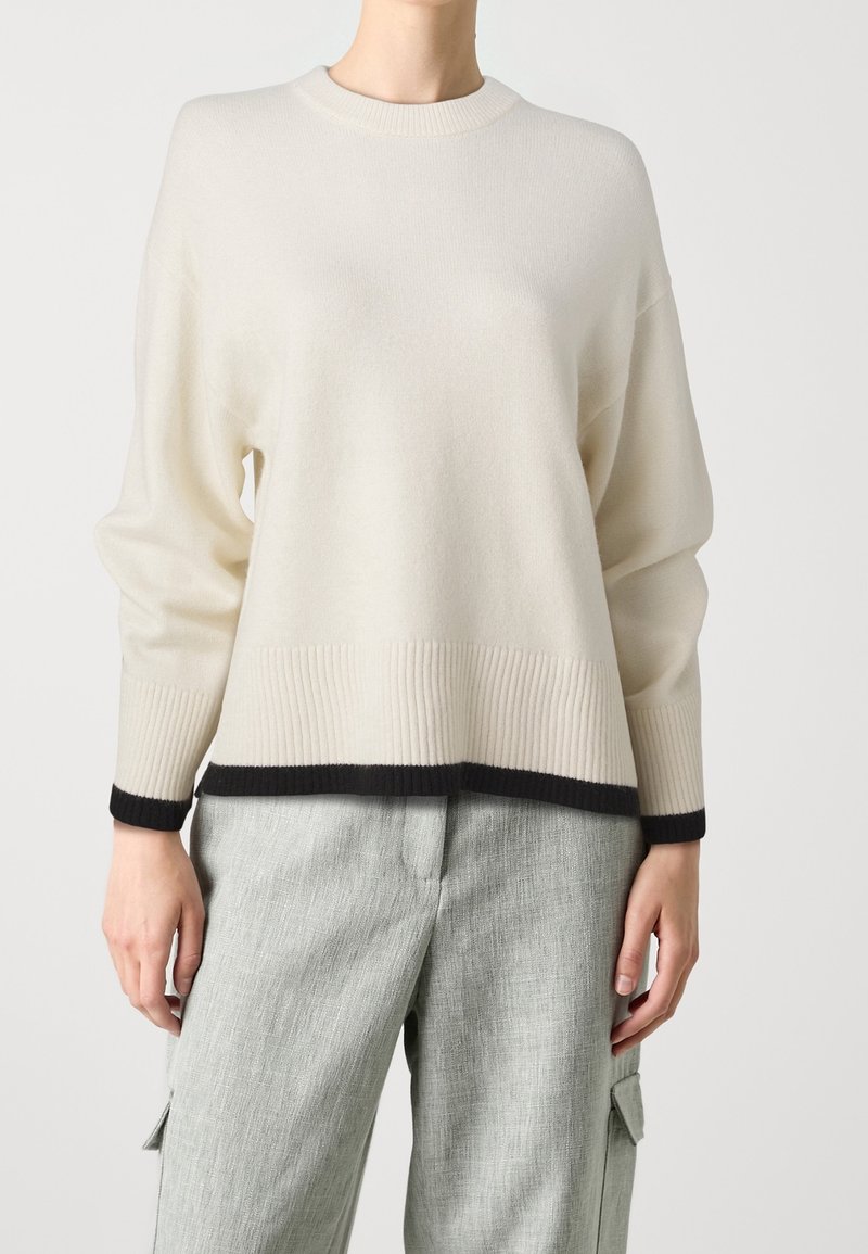 Personne portant un pull crème avec des bordures noires aux poignets et à l'ourlet, associé à un pantalon gris clair texturé avec des poches latérales.