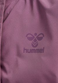 Lila Stoff mit einer strukturierten Oberfläche, einem eingeprägten "hummel"-Logo und einem dekorativen Emblem darüber. Das Design ist minimalistisch und modern.