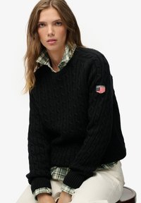 Zwarte kabelgebreide trui met geribbelde manchetten, gecombineerd met een groen-wit geruit overhemd. Voorzien van een logopatch op de mouw.