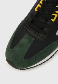 Mustad tenniskingad roheliste seemisnahast detailide, kollaste silmuste, mustade paelte ja tekstuursete materjalidega. Omavad valget tallad ja kummist mustriga tallapinda.