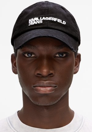 Mladi crnac nosi crnu kapu Karl Lagerfeld Jeans i bijelu košulju, gleda ravno s neutralnim izrazom lica.