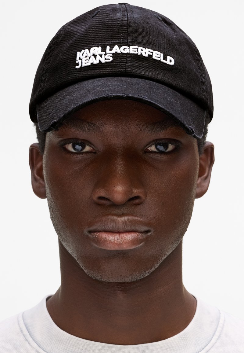 Νέος μαύρος άνδρας που φοράει μαύρο καπέλο Karl Lagerfeld Jeans και λευκό πουκάμισο, κοιτάζοντας μπροστά με ουδέτερη έκφραση.