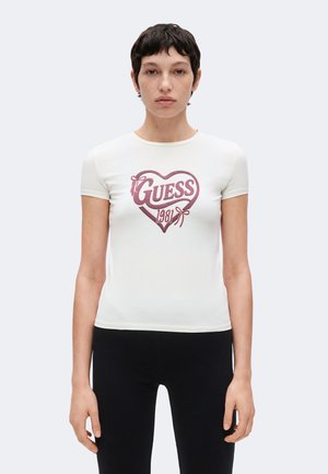 Femme aux cheveux courts et foncés portant un t-shirt blanc cintré avec un cœur rose et le texte "Guess 1981", associée à un pantalon noir, debout sur fond blanc.