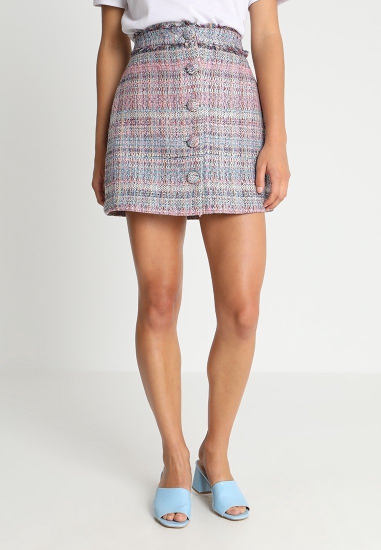 Femme portant une mini-jupe en tweed violet et bleu taille haute, boutonnée sur le devant, avec une ceinture froncée et des sandales à talons bleu clair.