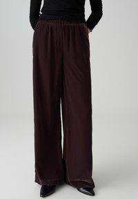 Wide-leg trousers in dark burgundy satin, featuring an elastic waistband and smooth texture, paired with black pointed-toe shoes.