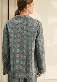 Dlouhý rukáv košile z tyrkysové látky s opakujícím se květinovým vzorem v růžové a bílé barvě. Má límeček a knoflíkové zapínání na zadní straně.