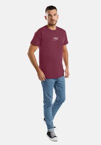 Burgunderfarbendes Baumwoll-T-Shirt mit kurzen Ärmeln, mit Logo-Details auf der Vorderseite, kombiniert mit hellblauen Jeans und schwarzen Sneakers. Lässiger Schnitt und Design.
