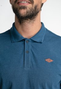 Camisa polo a rayas azules con una tapeta de dos botones, cuello acanalado y un distintivo logo naranja "PETROLIND" en el pecho.