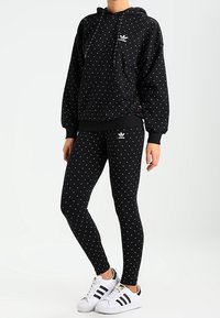 Person som bär en svart Adidas-hoodie och leggings med vitt prickmönster, kombinerat med vita Adidas-sneakers med svarta ränder.