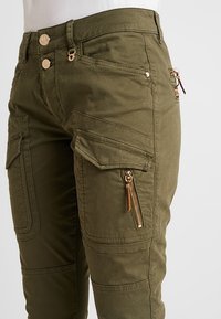 Fyrkantiga byxor i olivgrön bomullsblandning, med flera fickor med lock och dragkedjor, samt detaljer i guldton.