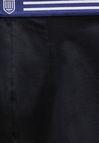 Κοντινή λήψη μαύρου υφάσματος με μπλε ζώνη μέσης που φέρει λευκές οριζόντιες ρίγες και ένα μικρό λευκό έμβλημα στην αριστερή πλευρά.