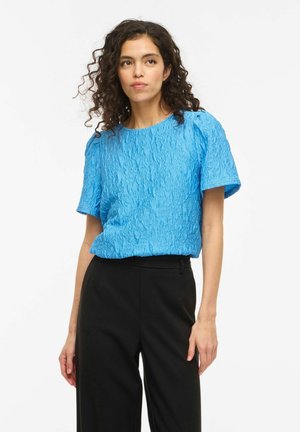 Kobieta o kręconych włosach, ubrana w teksturowaną, jasnoniebieską bluzkę z krótkim rękawem oraz czarne spodnie z wysokim stanem, stoi na białym tle.