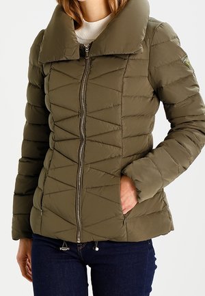Femme portant une doudoune matelassée vert olive avec fermeture éclair devant, debout avec une main dans la poche, vue des épaules aux hanches.