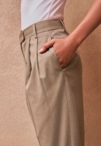 Pantaloni beige plissettati con un design elegante, tasca laterale, sottili righe verticali e una texture liscia.