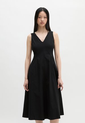 Femme aux longs cheveux noirs portant une robe midi noire sans manches avec un col en V, debout devant un fond clair uni.