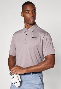 Svetlošedé polo tričko s čiernym logom Nike, s golierom a tromi gombíkmi, nosené s vzorovanými modrými šortkami a bielou golfovou rukavicou.