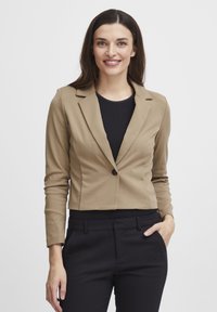 Kapad beige blazer med en enda svart knapp, släta tyg, slagkrage, bärs över en svart topp, ihop med svarta byxor.
