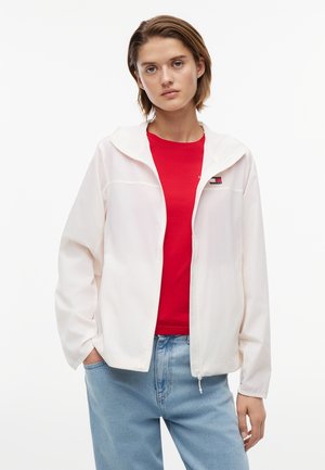 Νεαρός με κοντά μαλλιά που φοράει λευκό μπουφάν με φερμουάρ πάνω από κόκκινο πουκάμισο και ανοιχτόχρωμο μπλε τζιν, στέκεται με το ένα χέρι στην τσέπη.