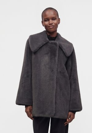 Personne aux cheveux courts portant un manteau ample en fausse fourrure gris foncé avec un large col et un pantalon noir sur un fond clair uni.
