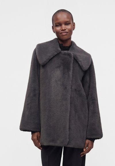 Personne aux cheveux courts portant un manteau ample en fausse fourrure gris foncé avec un large col et un pantalon noir sur un fond clair uni.