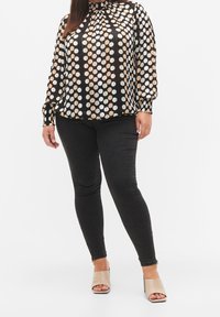 Blouse noire avec de grands pois irisés en blanc et nuances d'or, à manches longues, avec des fronces au niveau du cou, associée à un jean foncé et des mules beiges.