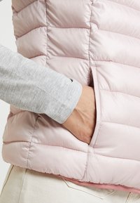 Hellrosa, gesteppte Steppweste mit seitlicher Tasche. Das Innere ist mit einem rosa Futter versehen und wird mit einer grauen Langarm-Shirt-Manschette kombiniert.