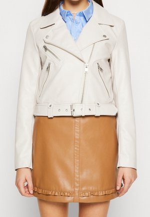 Femme portant une veste de motard blanche ceinturée sur une chemise bleue à boutons et une mini-jupe en cuir marron avec un volant.