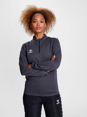 Žena s kudrnatými vlasy, která má na sobě tmavě šedý sportovní top s půlzipem a černé kalhoty s logy hummel, stojí se zkříženýma rukama na jednobarevném pozadí.