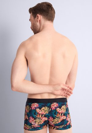 Homme aux cheveux bruns courts vu de dos, portant un boxer à motifs floraux, debout devant un fond clair uni.