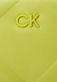Jasně žlutý materiál s texturou, na kterém je vyražené logo "CK". Povrch má geometrický vzor s diagonálními čarami vytvářejícími hloubku.