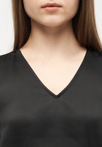 Γυναίκα με μακριά μαλλιά που φοράει μαύρο τοπ με V-neck, εμφανής από το πηγούνι έως το πάνω μέρος του στήθους με φόντο απλό.